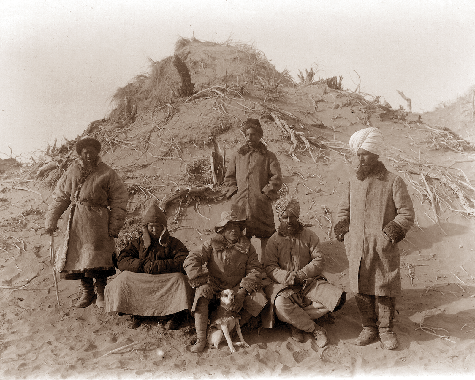 Photograph of Stein and assistants at Ulugh-Mazar in present-day China. Behind them is the rocky and desert landscape of Central Asia.